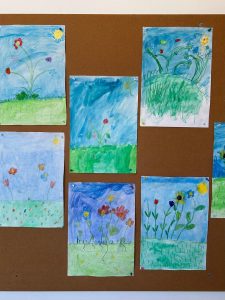 Mehrere bunte Kinderzeichnungen von Blumen auf einer Pinnwand, mit blauem Himmel und grüner Wiese als Hintergrund.