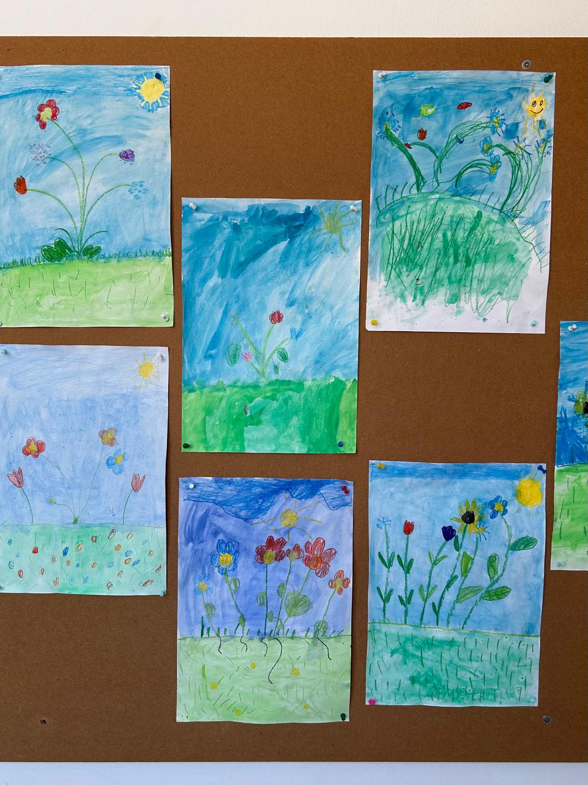 Mehrere bunte Kinderzeichnungen von Blumen auf einer Pinnwand, mit blauem Himmel und grüner Wiese als Hintergrund.