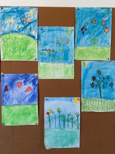 Kinderbilder mit Blumen und Wiesenlandschaften hängen auf einer Pinnwand, gemalt mit kräftigen Farben und einfachen Formen.