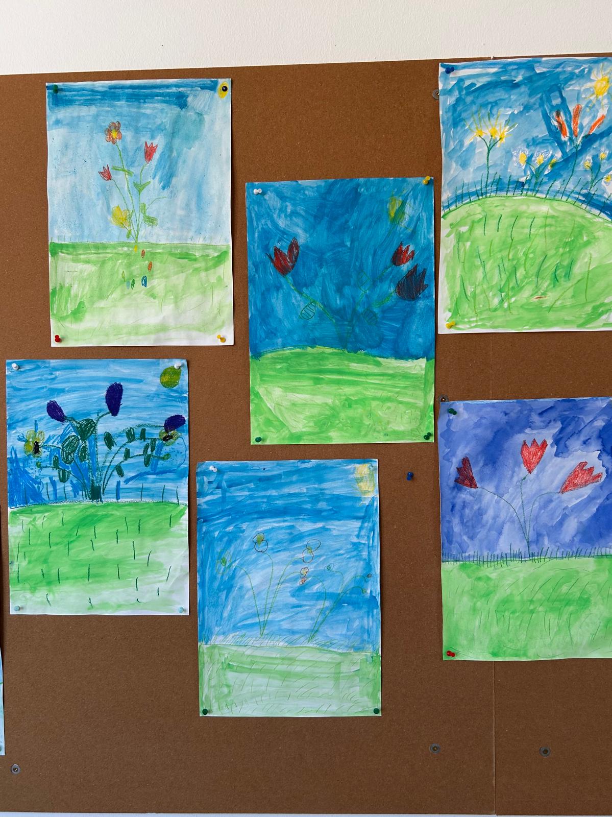 Verschiedene gemalte Blumenbilder von Kindern, befestigt an einer braunen Pinnwand, mit viel Blau und Grün im Motiv.