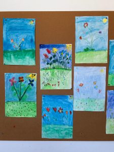 Kinderzeichnungen von Blumen auf einer Pinnwand, mit unterschiedlichen Blütenformen, leuchtenden Farben und sonnigen Himmelsszenen.